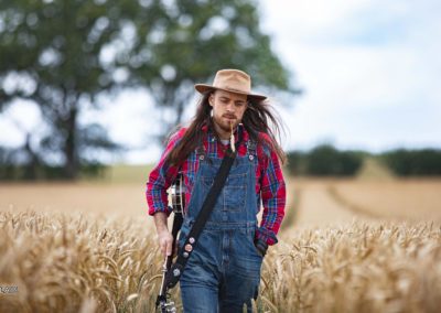 Portrait d'un joueur de banjo dans un champ de blé, portant un chapeau, une chemise rouge et une salopette bleu. Il marche. Style Country, Americana. Raphaël Fourage, à Abbaretz