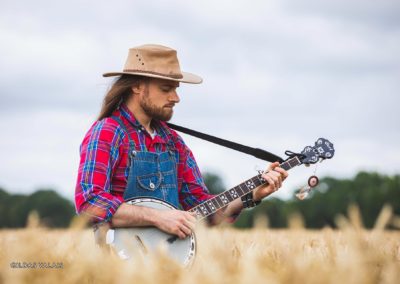 Portrait d'un joueur de banjo dans un champ de blé, portant un chapeau, une chemise rouge et une salopette bleu. Style Country, Americana. Raphaël Fourage, à Abbaretz